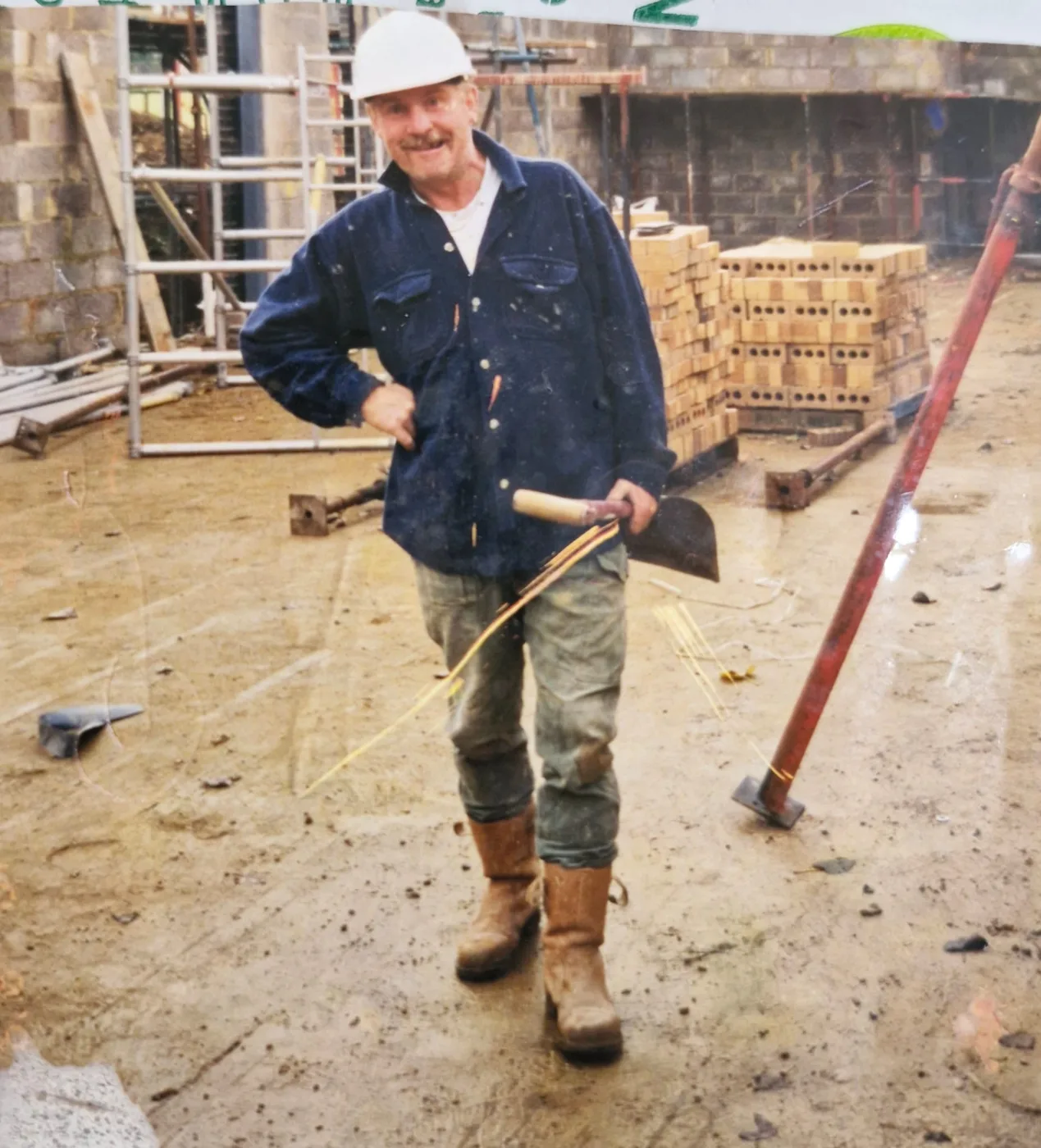 Tony Williams in a white hard hat, work jacket and boots, holding a pickaxe on a building site with scaffolding and stacked bricks behind him
