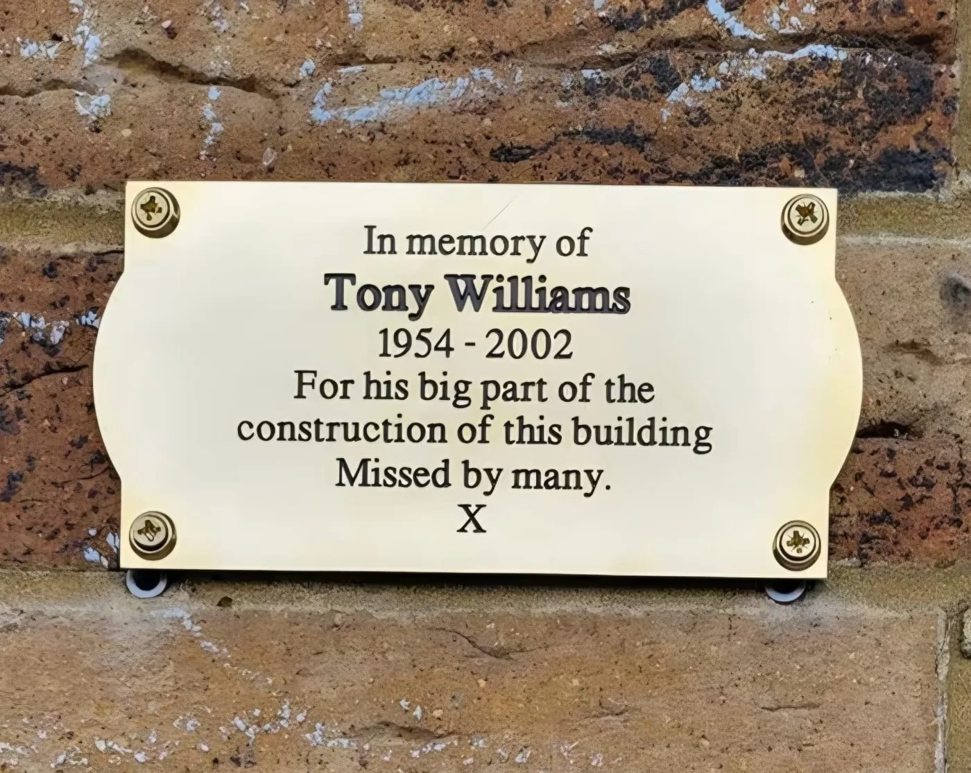 A brass plaque on a brick wall reading: In memory of Tony Williams, 1954 to 2002, for his big part of the construction of this building, missed by many